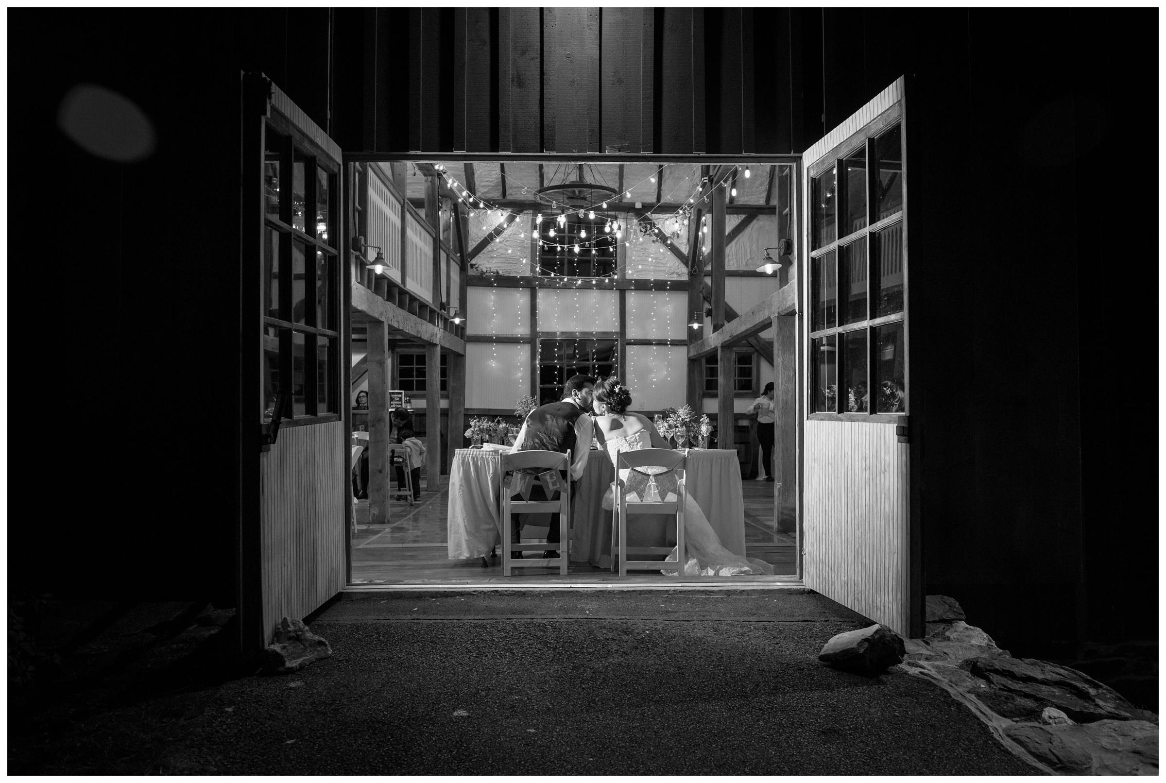 Bride and groom kiss in open barn doorframe during rustic wedding reception.