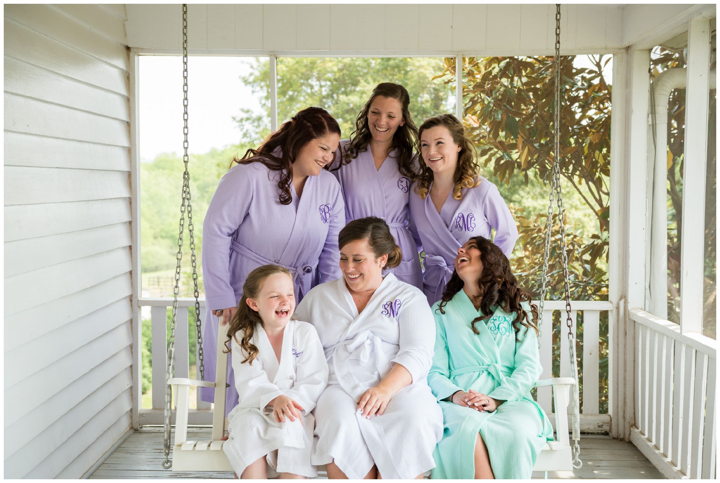 bride and bridesmaid laughing on farmhouse porch swing wearing matching robes