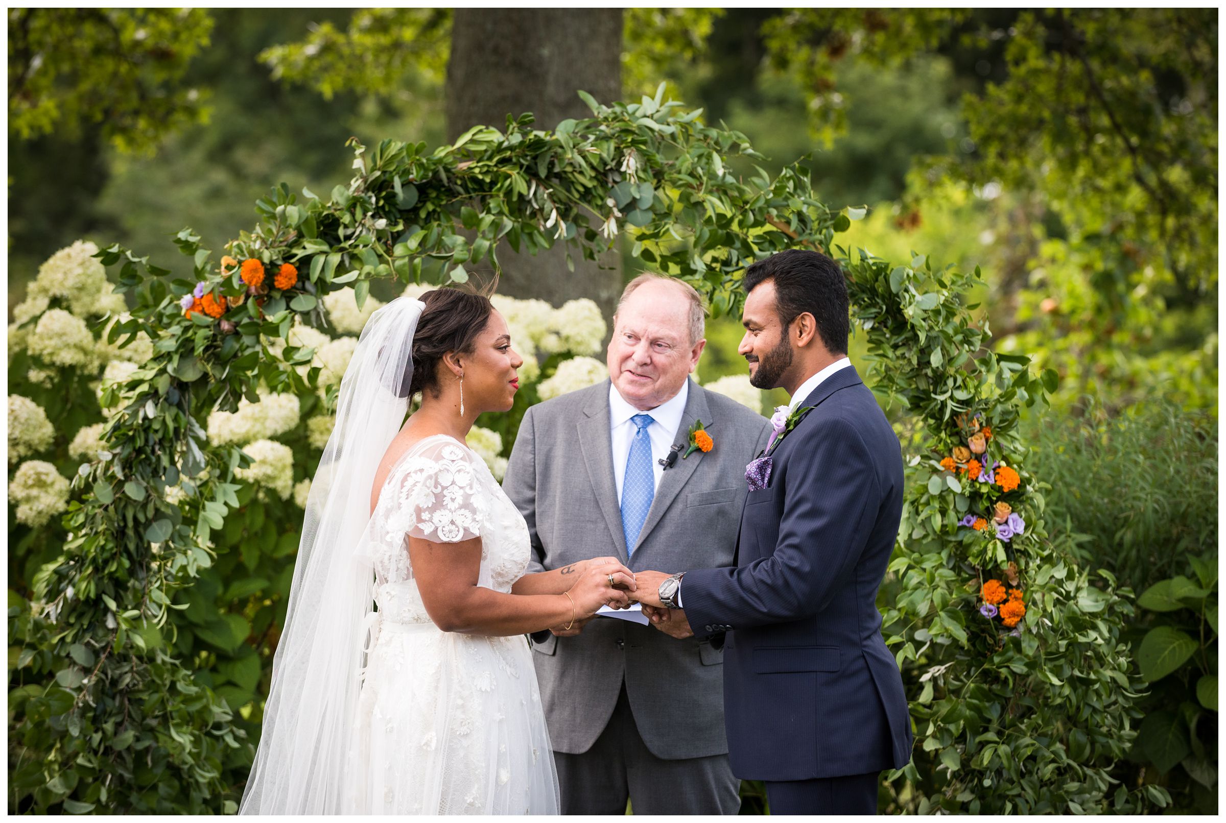 bride an groom exchange ring during summer wedding in front of floral arch with marigolds at Jorgensen Farms