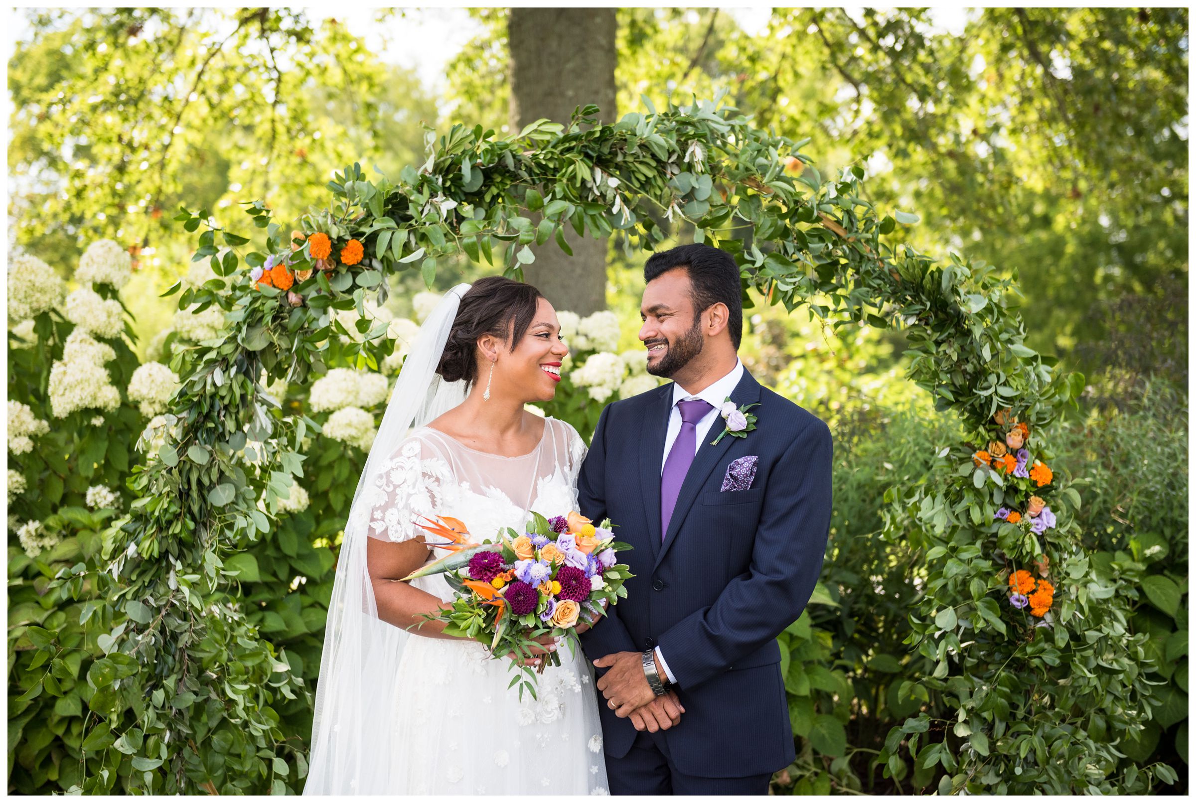 Columbus wedding photography of black bride and Indian groom under floral moongate arch with colorful purple and orange flowers