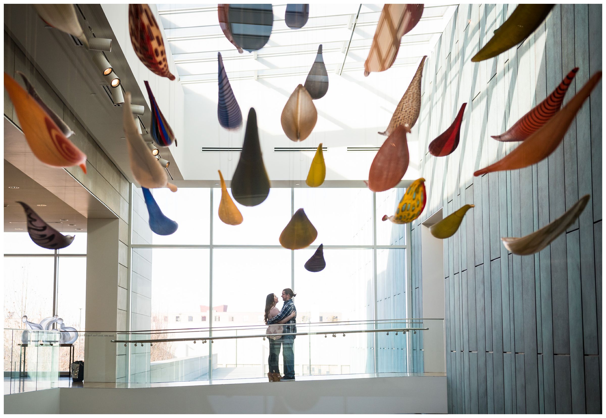 Engaged couple standing in the atrium of the Columbus Museum of Art surrounded by colorful hanging glass artwork.