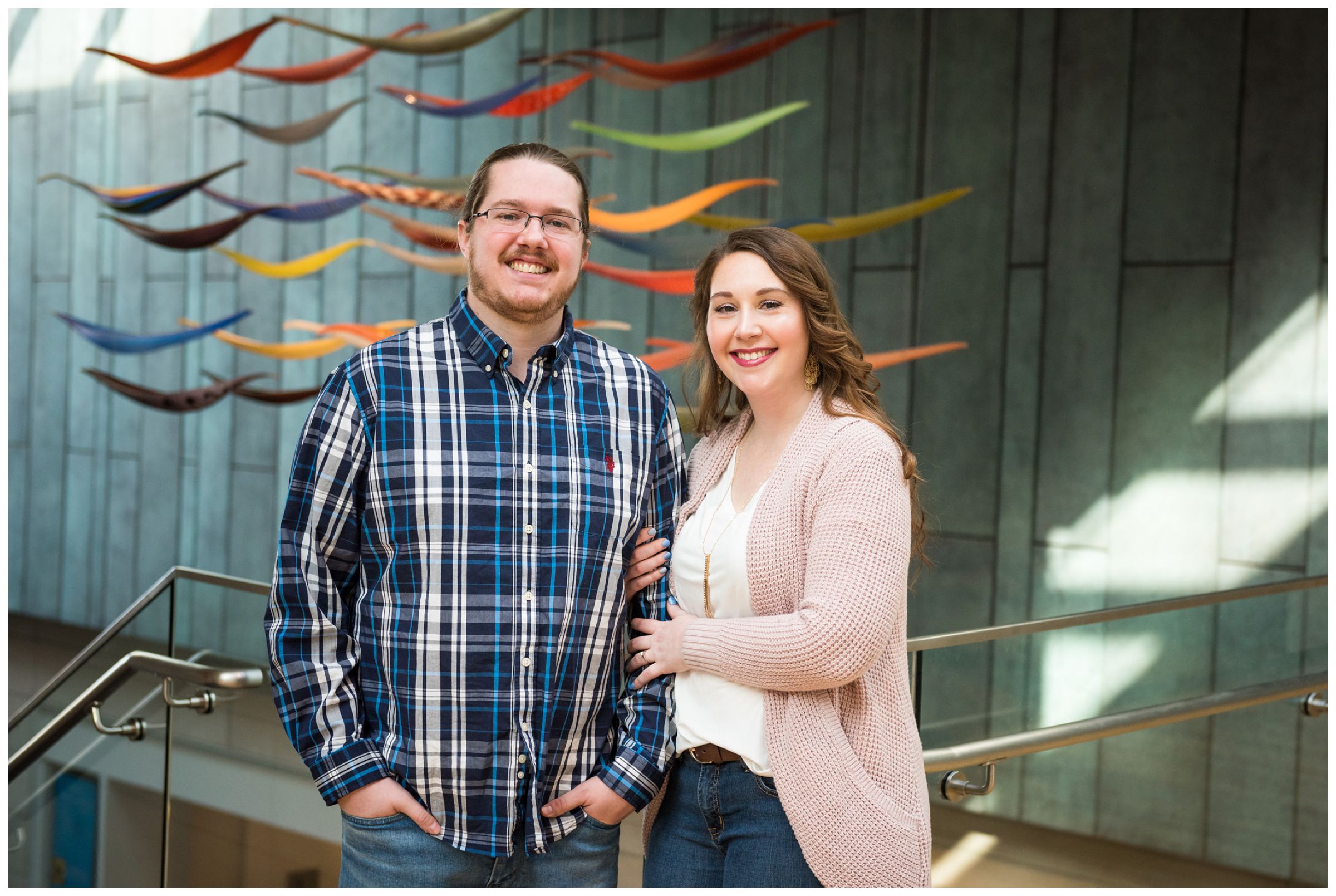 Winter indoor engagement photos in the atrium of the Columbus Museum of Art surrounded by colorful hanging glass artwork.